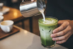 A close-up, dynamic shot of a barista's hands pouring intricate latte art with steamed milk into a glass of iced matcha latte. The layers of green and white are distinct. Cafe ambiance. Bokeh background. Highly detailed, food photography style.
