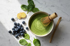 A vibrant, top-down shot of a whisked bowl of matcha, surrounded by healthy ingredients like blueberries, spinach, and ginger on a clean white marble background. The lighting is bright and natural. Photorealistic, high-detail.